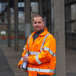 Eine Person steht am Bahnhof und trägt eine orange Warnjacke und einen Ordner.