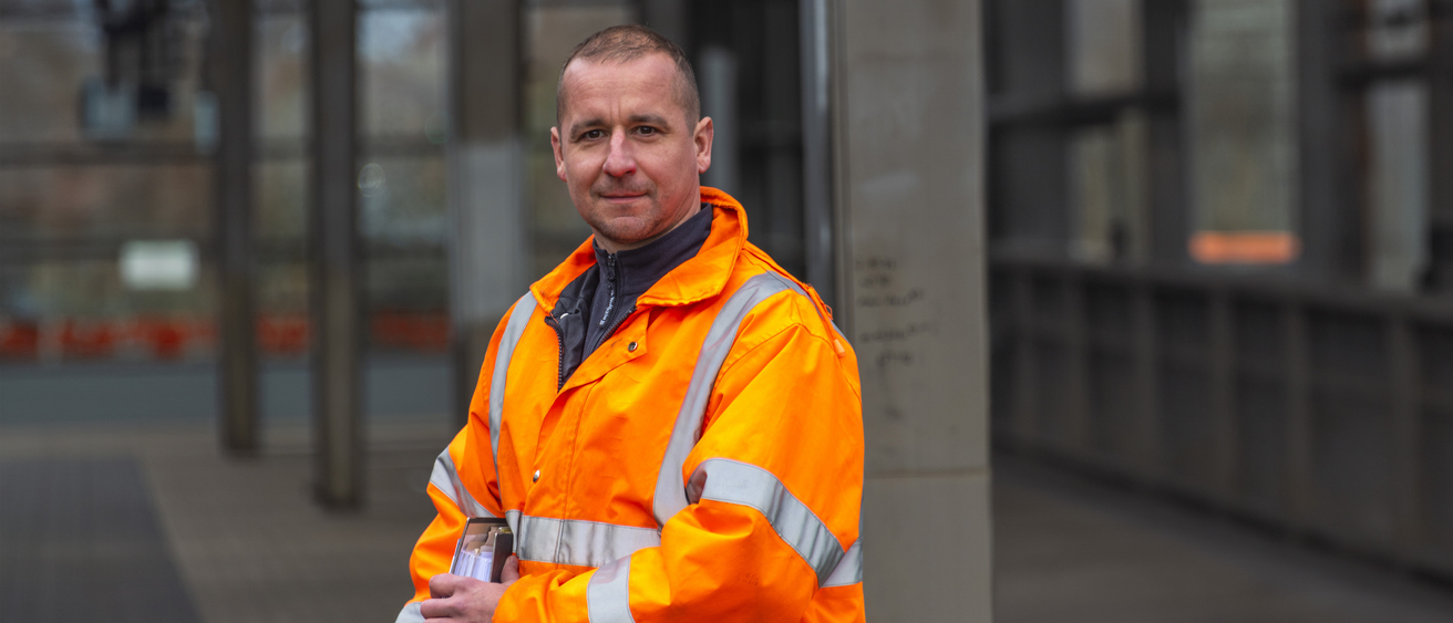 Eine Person steht am Bahnhof und trägt eine orange Warnjacke und einen Ordner.